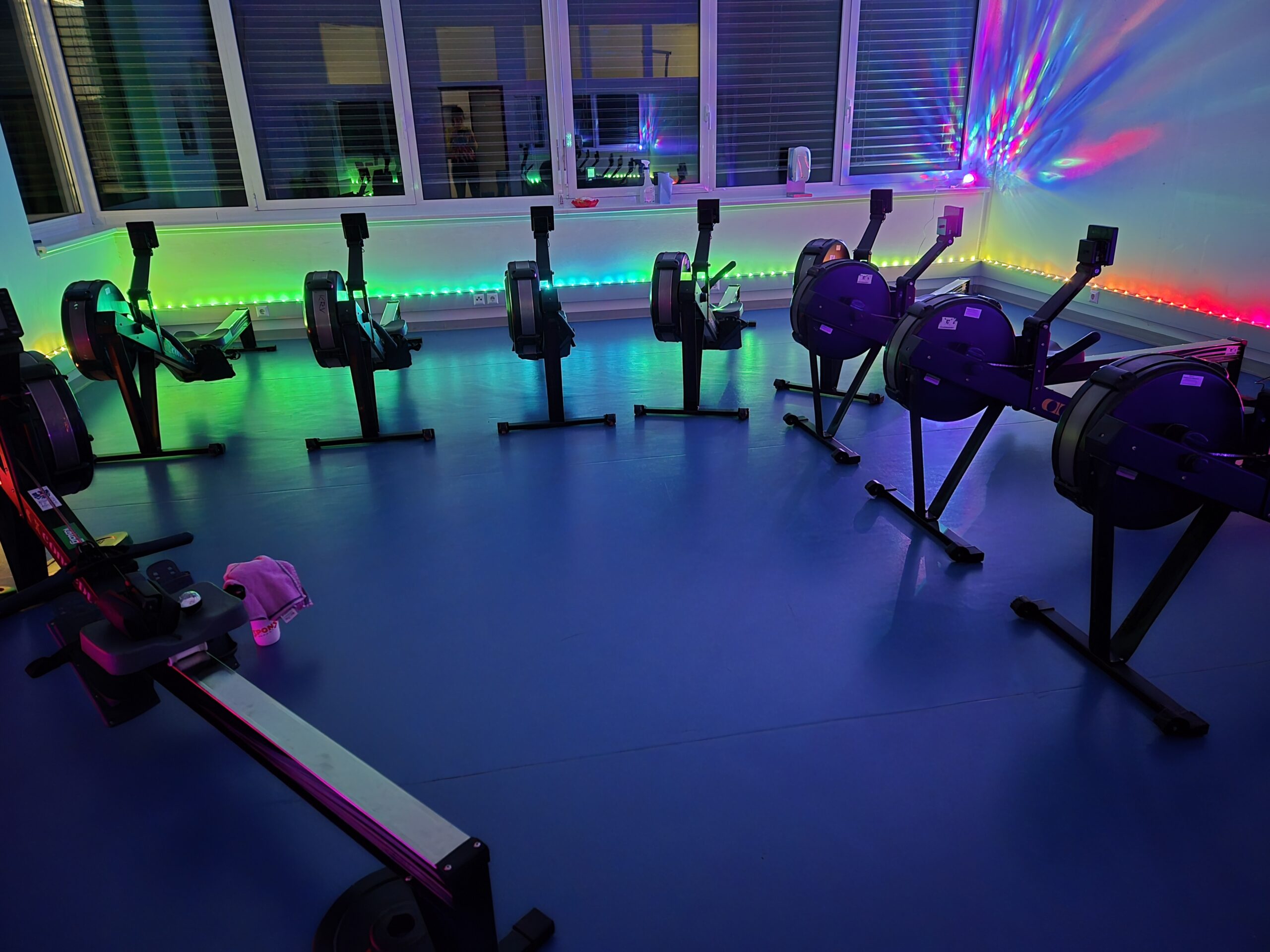 Mehrere Ruderergometer in einem abgedunkelten Raum mit bunten LED-Lichtern an den Wänden. Die Geräte sind kreisförmig angeordnet, während farbige Lichter in Grün, Blau und Pink eine lebhafte, clubartige Atmosphäre erzeugen.