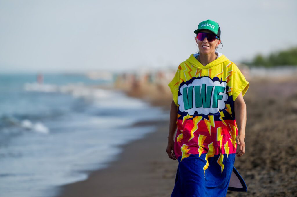Ruderin Magdalena Lobnig spaziert am Strand entlang, trägt eine bunte Ponchojacke mit der Aufschrift „VWF“ und eine grüne Kappe mit der Aufschrift „Kelag“.