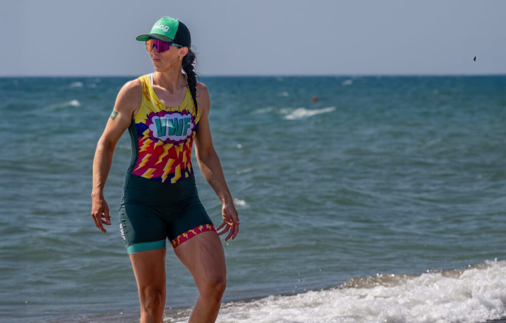 Magdalena Lobnig steht nach dem Rennen im seichten Wasser am Meer, trägt einen farbenfrohen Ruderanzug mit „VWF“-Logo und eine grüne Kelag-Kappe.